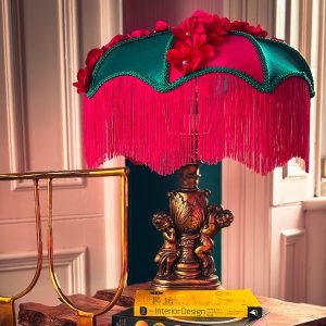 A green and pink lampshade with a deep fringe is styled on a table.
